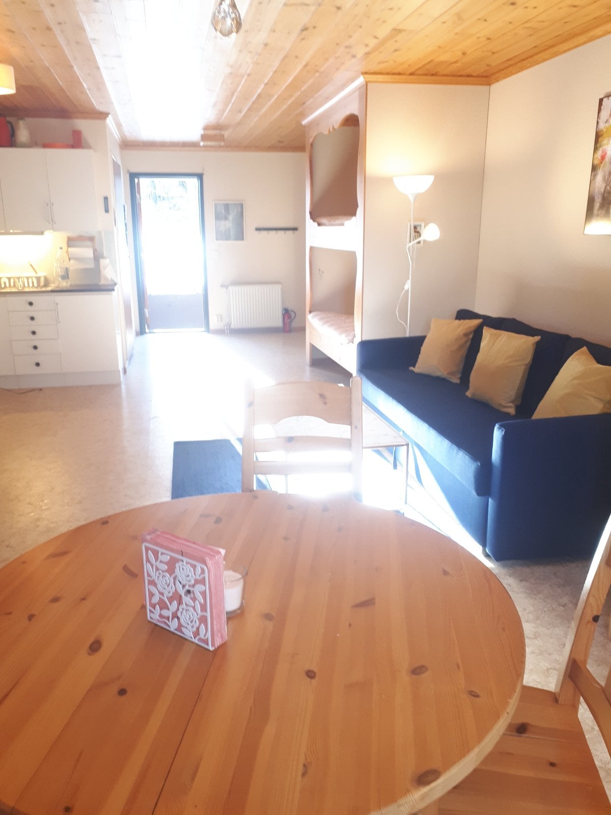 An open living area features a wooden dining table with a small decorative item, complemented by a blue sofa adorned with yellow cushions. Natural light floods the space through large windows, enhancing the wood-paneled ceiling and the bright, welcoming atmosphere.