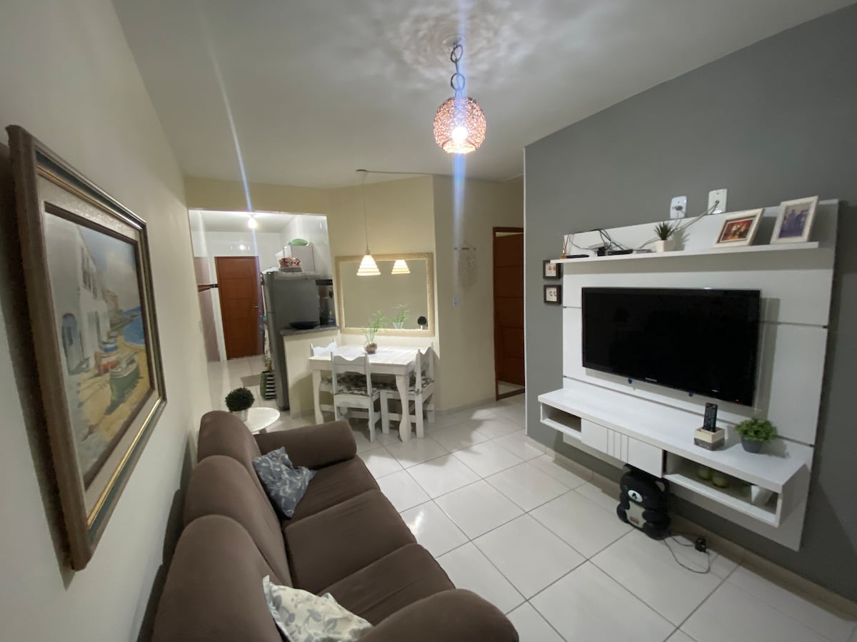 An inviting living area is presented with a comfortable brown sofa positioned beside a contemporary TV unit. A dining table with four white chairs is visible in the background. Soft lighting from a pendant fixture creates a cozy ambiance, complemented by decorative wall art.
