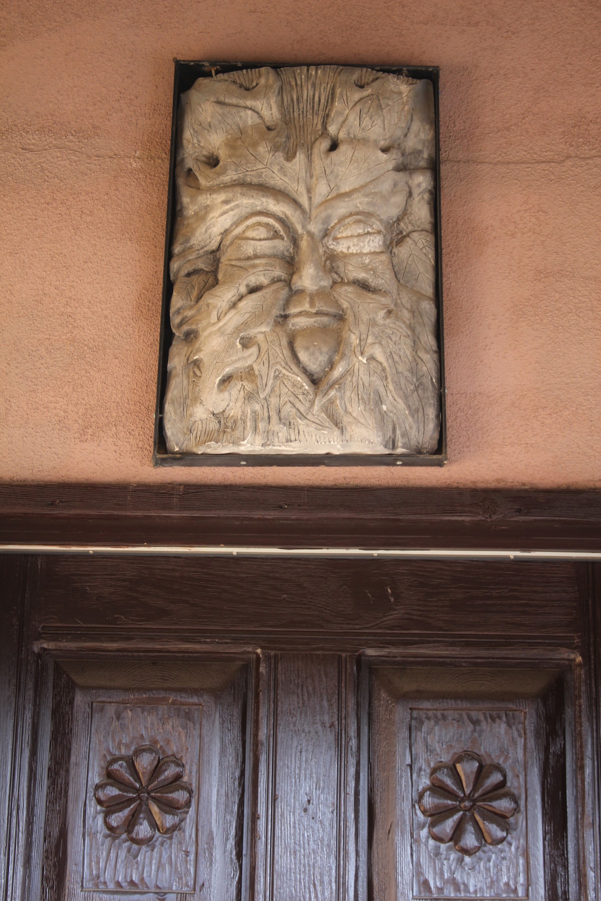 A hand-carved relief of a serene face, surrounded by stylized foliage, is mounted above a wooden door. The door features intricate floral carvings, adding a touch of craftsmanship to the entrance. The warm tones of the surrounding wall complement the earthy aesthetics.