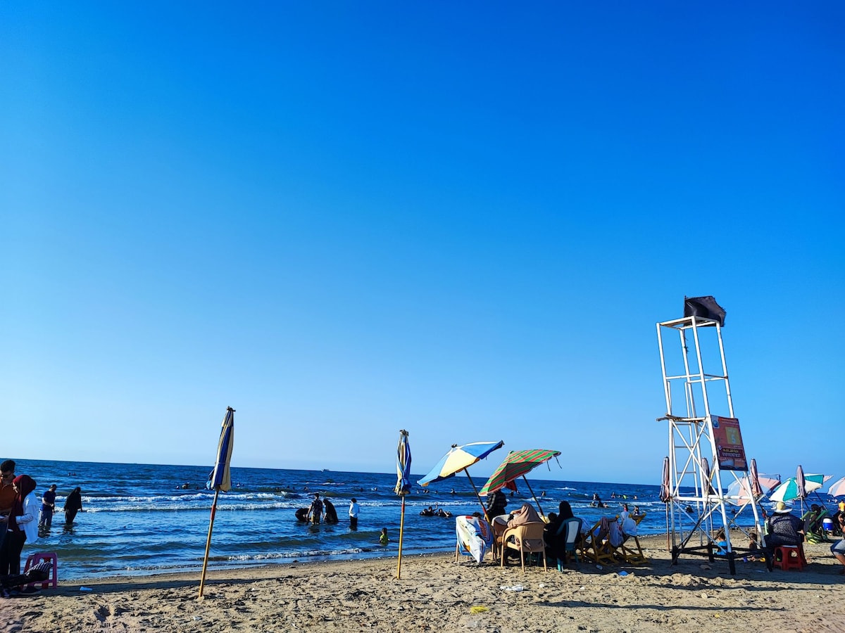A sandy beach scene is depicted, featuring umbrellas and beach chairs arranged along the shoreline. Individuals are seen enjoying the water, while a lifeguard tower stands in the background. The clear blue sky creates a bright atmosphere over the tranquil ocean waves.