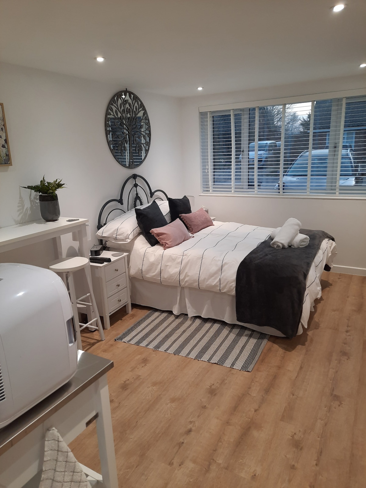 A bright and contemporary bedroom features a spacious bed adorned with decorative pillows and a throw. A mirror reflects natural light from the window, complemented by neutral-toned walls. A small seating area and kitchenette are visible, enhancing the room's functionality.
