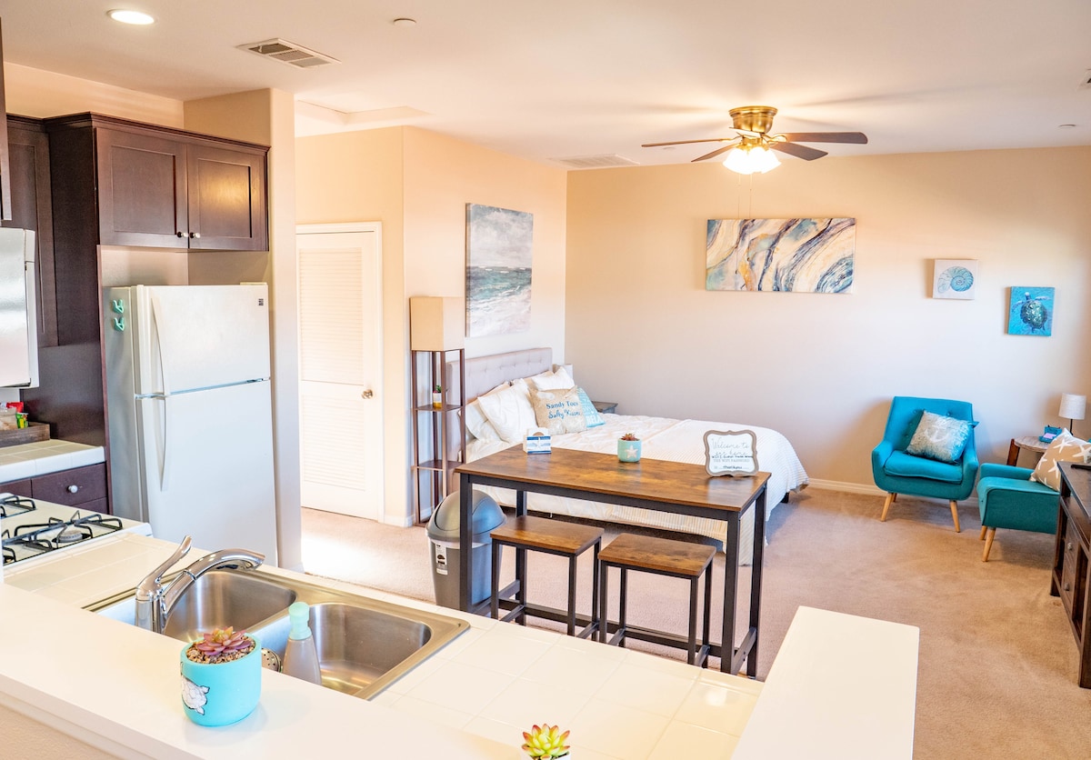 A bright and airy studio features an open layout with a full kitchen on one side and a cozy sleeping area on the other. A central dining table is accompanied by chairs, and a small sitting area includes a blue armchair. Natural light fills the room.