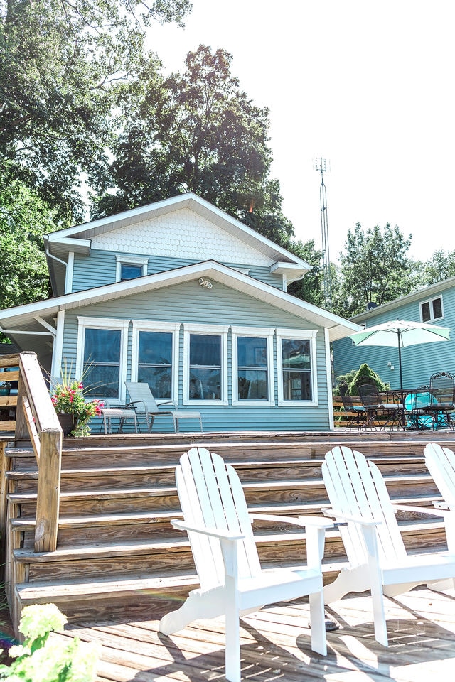The exterior of Three Sisters Cottage features a charming façade with light blue siding and a welcoming porch. Five white Adirondack chairs are positioned on a wooden deck, overlooking a serene outdoor space lined with greenery. Large windows provide views of the surroundings.