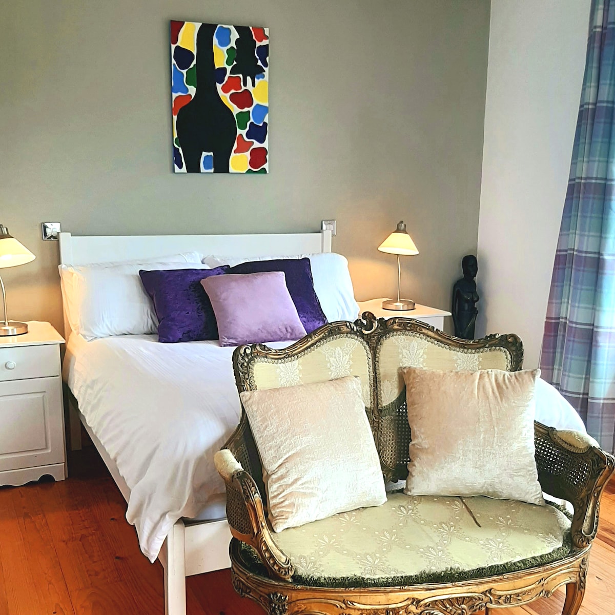 A well-appointed bedroom features a spacious bed with crisp white sheets and decorative purple pillows. Two bedside lamps provide soft lighting. An elegant seating area with a vintage-style loveseat complements the room, while a colorful artwork adorns the wall, enhancing the overall aesthetic.