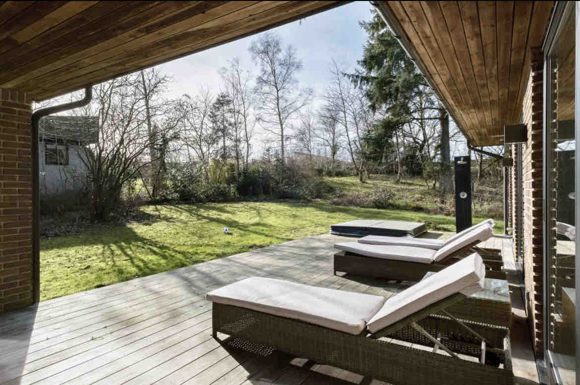 A spacious wooden terrace features two lounge chairs positioned for relaxation under natural sunlight. Lush greenery extends across the background, complemented by mature trees. A hot tub is visible, inviting quiet moments outdoors.