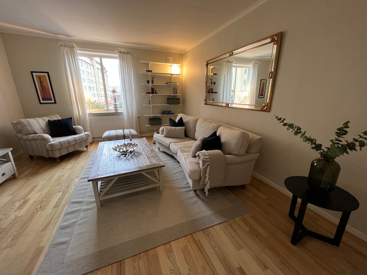 A cozy living area is featured, showcasing a light-colored sofa accompanied by a striped armchair. A coffee table sits at the center, while a large mirror reflects natural light from the window. Shelves display decorative items, enhancing the inviting atmosphere.