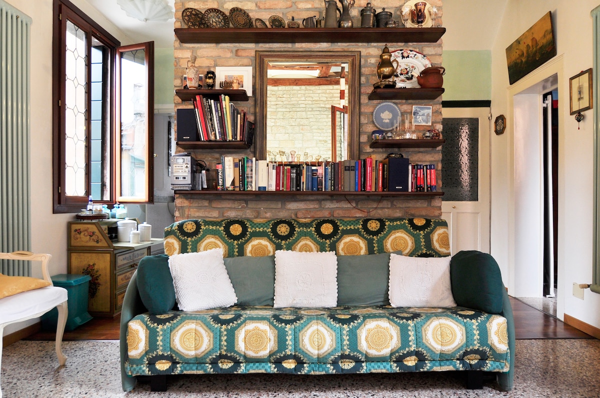 A cozy sitting area features a patterned sofa adorned with white cushions. A stone wall serves as a backdrop, showcasing an assortment of books and decorative items on a wooden shelf. Natural light enters through the nearby windows, enhancing the warm ambiance of the space.