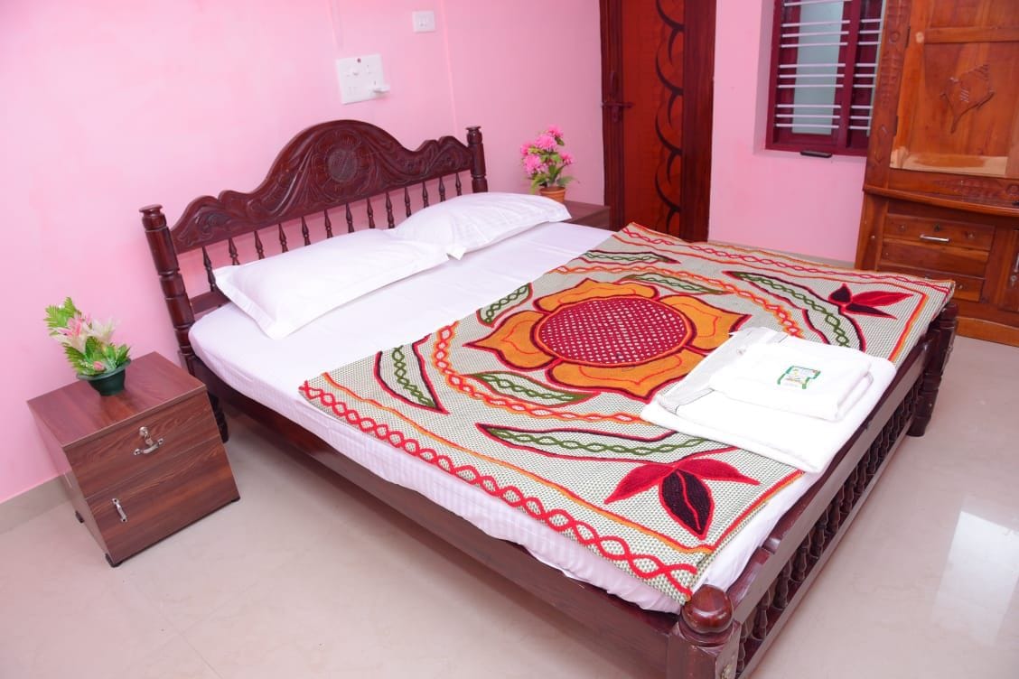 A bright bedroom features a wooden bed with a decorative patterned blanket draped over it. Two clean white pillows sit atop the spacious bed. A bedside table holds a small potted plant, and a wooden wardrobe is visible in the background, against soft pink walls.