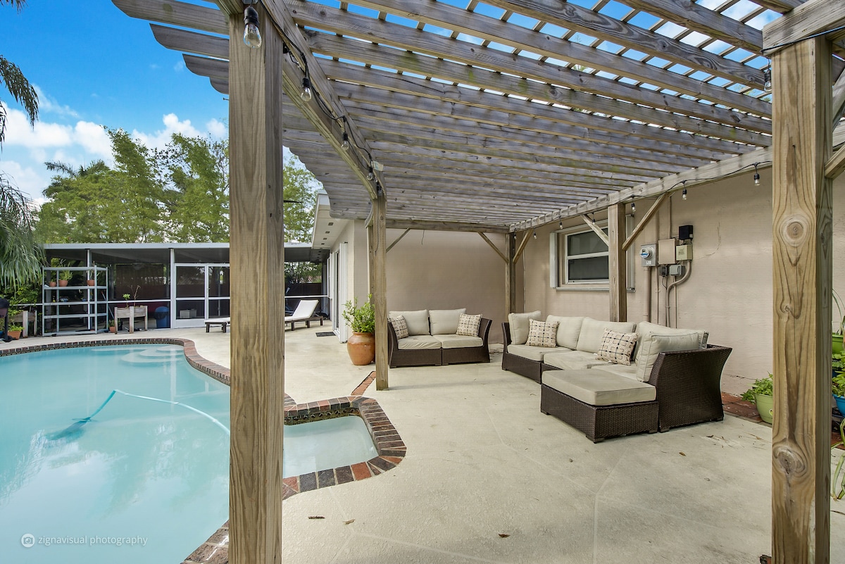 A covered patio area features comfortable seating arranged around a coffee table, providing a shaded space for relaxation. The adjoining swimming pool reflects the surrounding greenery, while a light breeze is invited through the open design of the space.