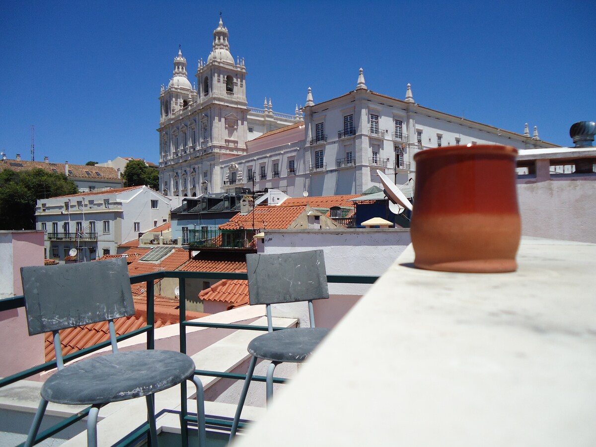 A rooftop terrace is displayed, featuring two metal chairs positioned near a stone railing. Terracotta pottery rests on the surface, with a backdrop of Lisbon's rooftops and historical buildings under a clear blue sky.