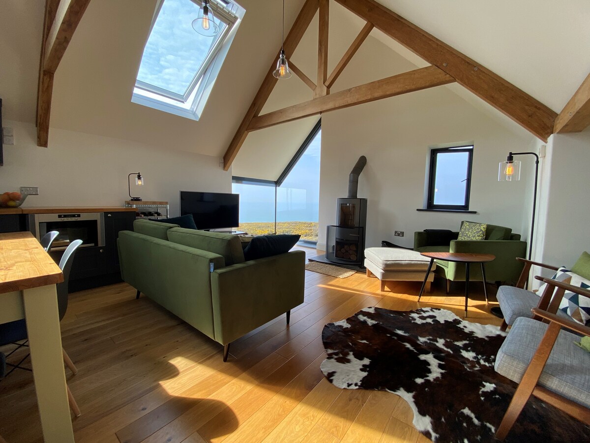 An open-plan living area features large windows offering sea views. A modern kitchen is paired with a dining table. Green sofas and armchairs create a comfortable seating arrangement, complemented by a log-burning stove. Exposed wooden beams add character to the high ceiling.