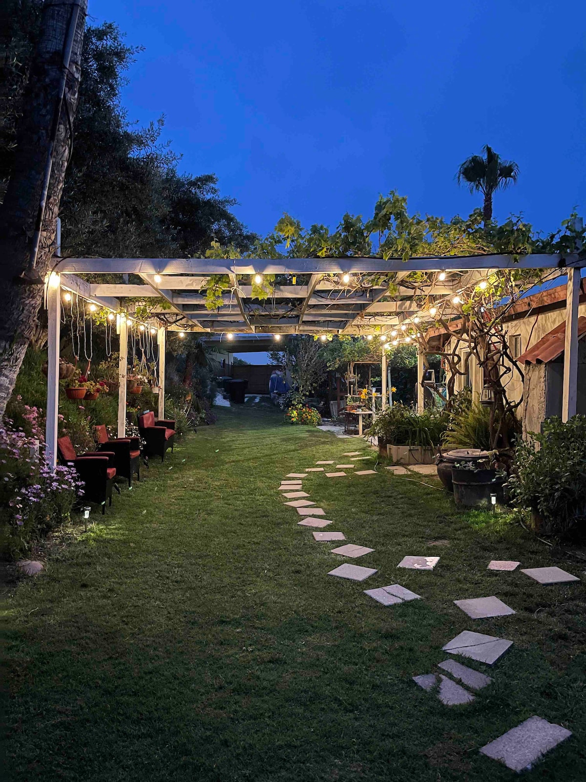 A garden path, lined with stepping stones, leads through a landscaped area illuminated by string lights. A pergola provides shade, while seating areas are arranged under the greenery. Flowerbeds add color to the space, creating a serene outdoor environment.