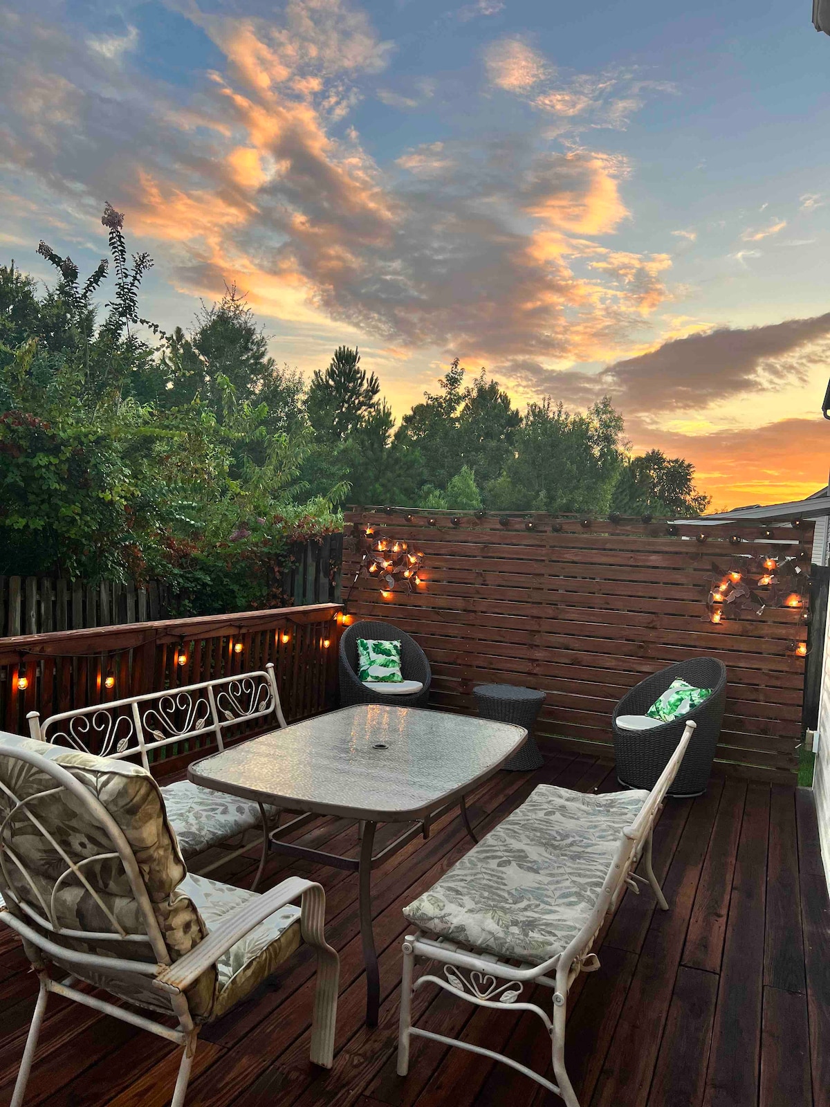 A back patio is depicted, featuring a rectangular table surrounded by chairs. The area is enhanced by string lights, creating a gentle glow against a backdrop of lush greenery. A colorful sunset is visible in the sky, adding warmth to the setting.