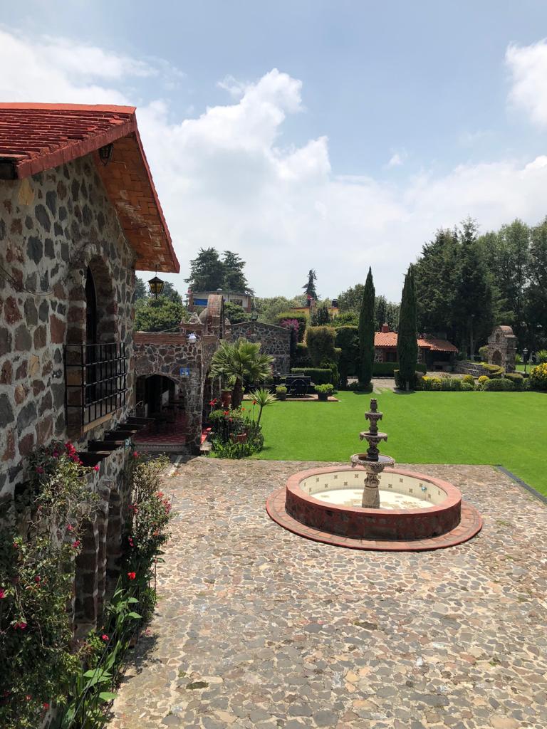 A scenic view features a stone courtyard with a circular fountain at the center. Lush green lawns and carefully maintained gardens surround the area, complemented by blooming flowers and tall trees. The rustic architecture of the nearby buildings is accentuated by red-tiled roofs.