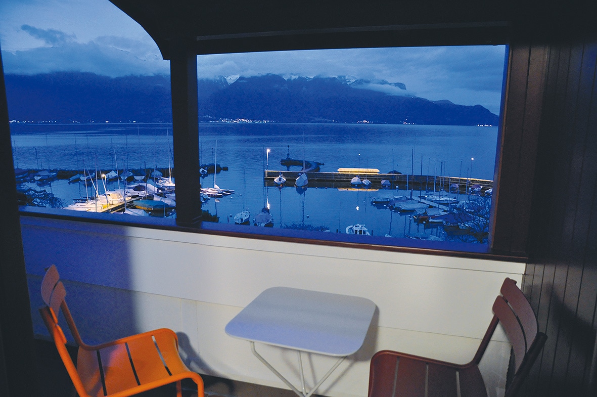 A balcony with two chairs and a small table offers a serene view of the lake and snowy mountains beyond. The scene is illuminated with soft lights reflecting off the water and boats moored at the nearby marina, creating a tranquil evening atmosphere.