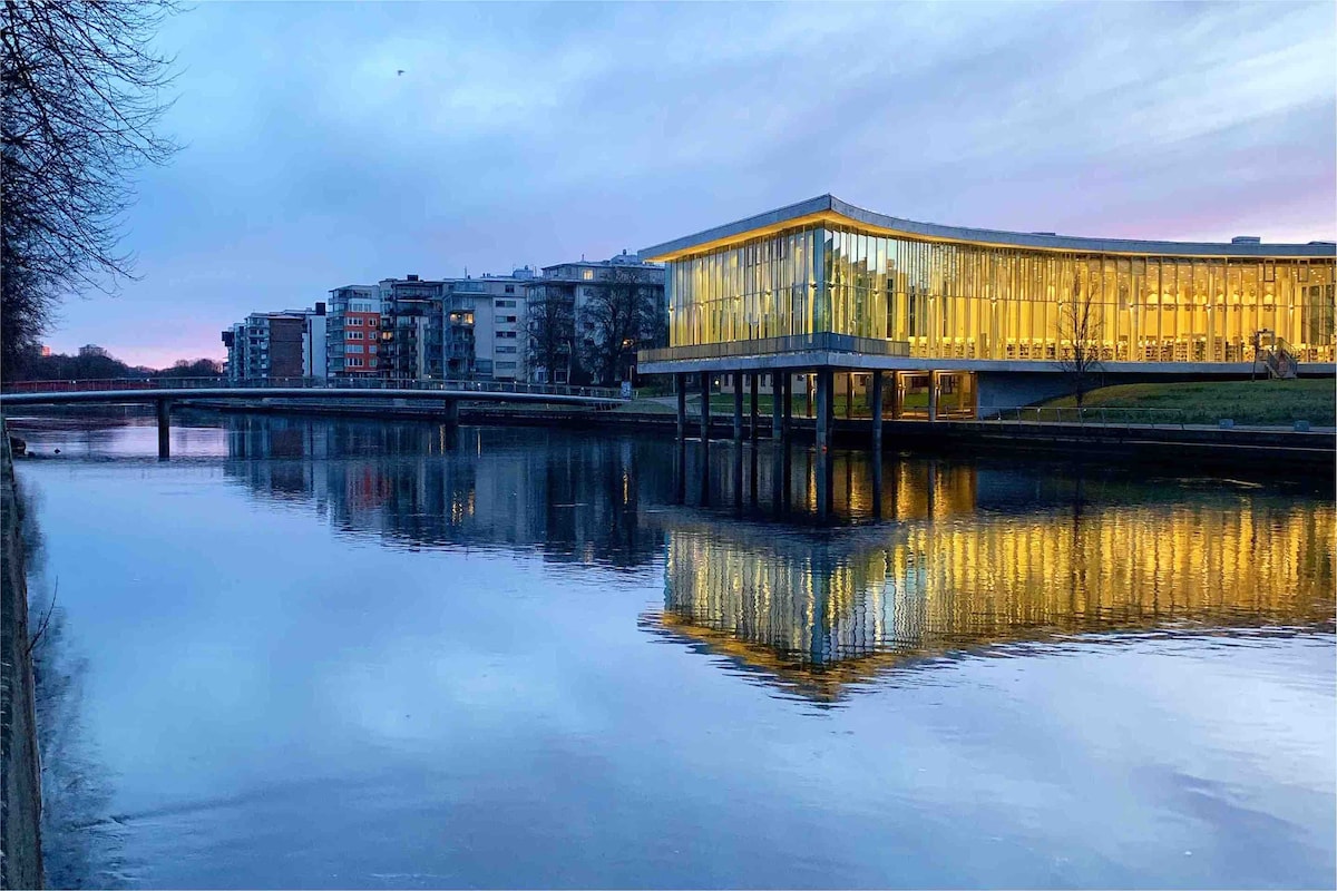 A modern building with a sleek glass facade reflects softly in the river at dusk. The structure is elevated on stilts, surrounded by a tranquil waterway, while nearby residential buildings provide a contrasting backdrop. The sky transitions from blue to soft purple hues.