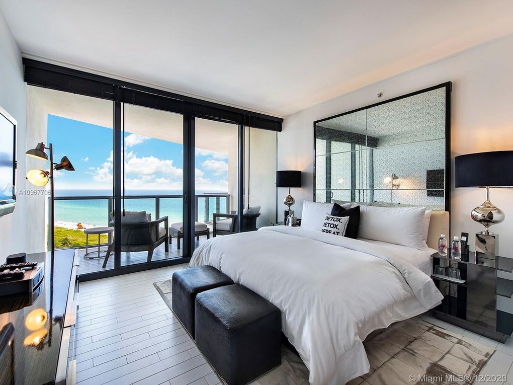 A modern bedroom features a spacious bed with crisp white linens, flanked by stylish black bedside tables with lamps. Large glass doors offer a view of the Atlantic Ocean, allowing natural light to brighten the room. A sleek dresser and minimalist decor complete the inviting atmosphere.