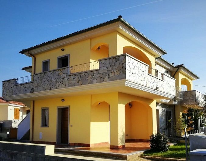 A two-story house is shown, featuring a sunny yellow exterior and stone accents. Balconies are present on both levels, with arched openings. A patio area is visible, along with landscaped greenery in the foreground and a clear blue sky above.