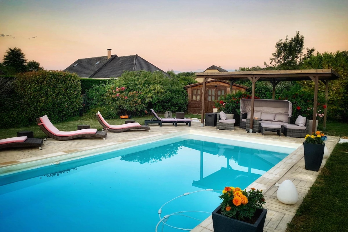 A serene outdoor space features a swimming pool surrounded by lounge chairs and shaded seating. Lush greenery and flowering plants enhance the setting, while a wooden gazebo offers additional seating. The area is tranquil, with a soft evening light reflecting off the pool's surface.