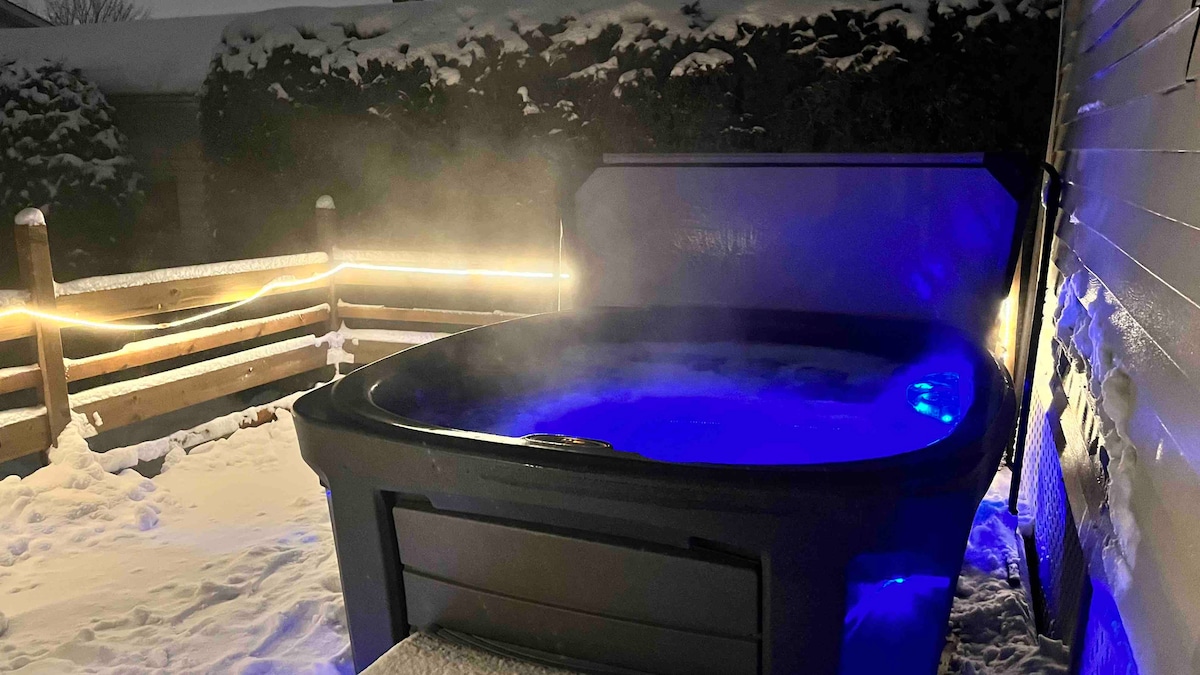 A hot tub is set against a snowy backdrop, gently steaming in the cold air. Soft blue lighting highlights the water, while surrounding foliage is partially obscured by snow. Warm light from nearby features creates an inviting atmosphere in the tranquil outdoor space.