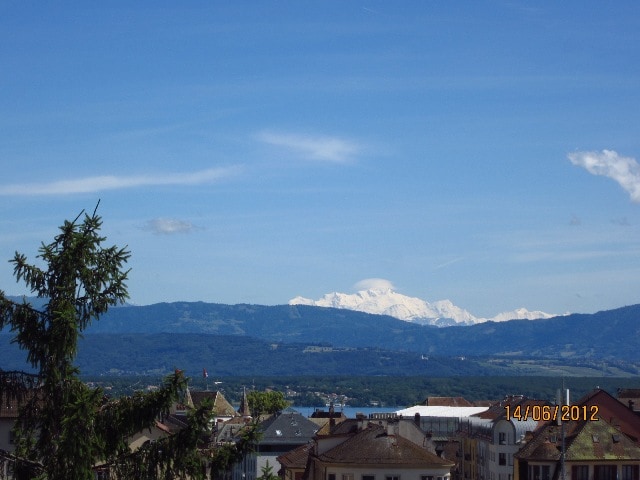 A clear view of the snow-capped Alps is presented, with the expansive landscape of rolling hills and a serene lake in the foreground. A mix of rooftops is visible, adding a charming urban element to the scene. The sky is bright and blue, with a few clouds.