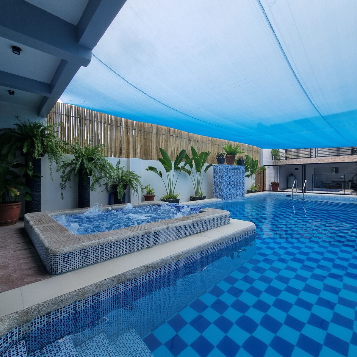 A swimming pool area is featured, adorned with blue tiles and surrounded by tropical plants. A raised hot tub with jets complements the pool, while a canopy provides shade above. The tranquil space invites relaxation in a well-maintained environment.
