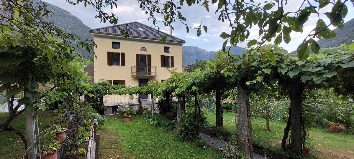 The exterior of the apartment is framed by a lush garden featuring grapevines and various greenery. A two-story building with a balcony is visible, set against a backdrop of mountains, creating a serene outdoor space. The lawn is well-maintained, enhancing the natural surroundings.