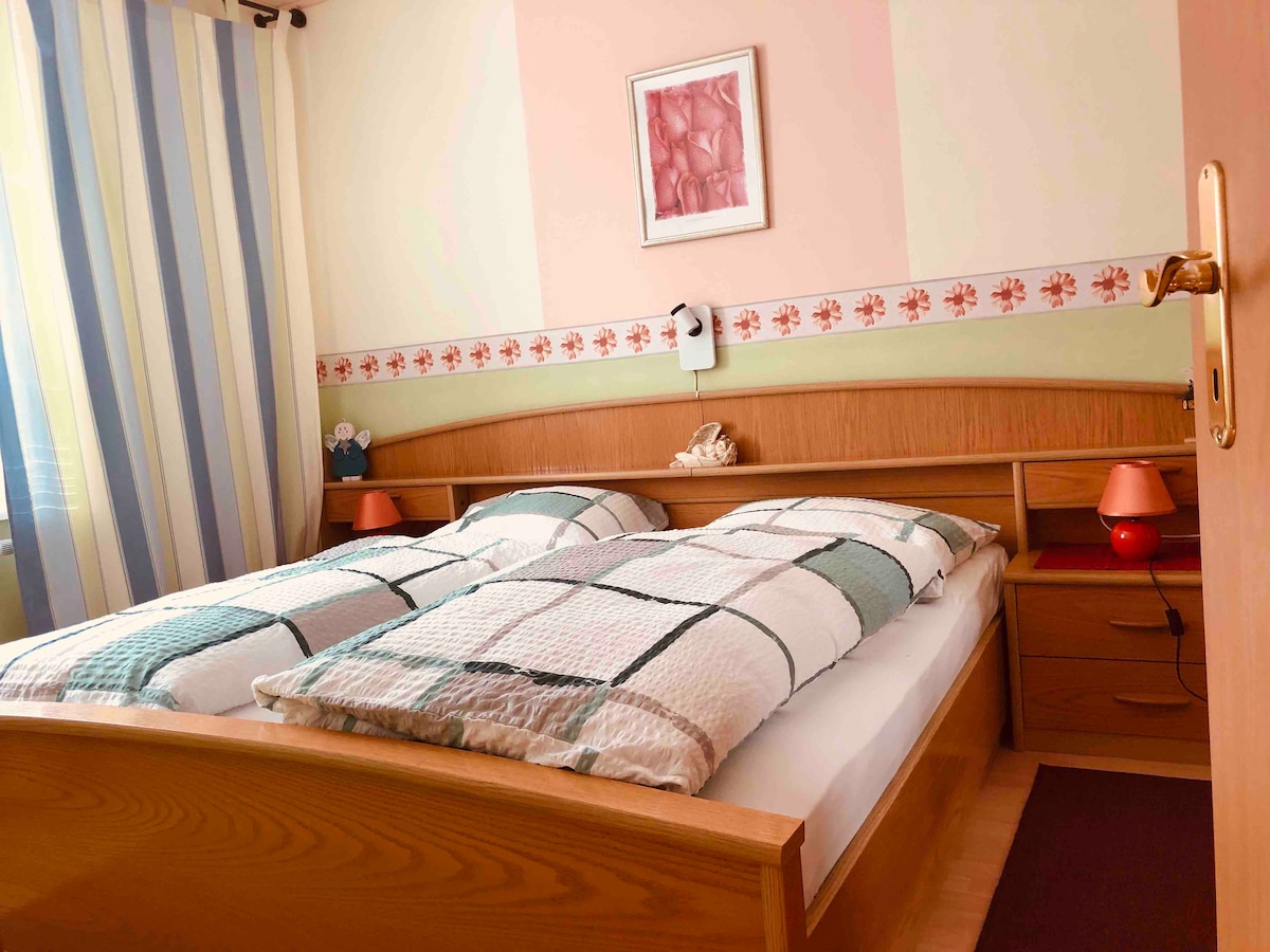 A well-lit bedroom features two individual beds covered with a checked quilt. Soft lighting is provided by bedside lamps, and a small decorative piece is displayed on the headboard. Colorful curtains frame the window, enhancing the cozy atmosphere.