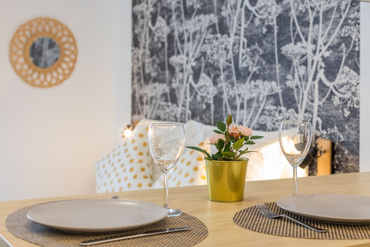 A dining area is presented with two plates and wine glasses, complemented by a small potted plant. A patterned wallpaper featuring delicate tree designs creates a serene backdrop. A mirror with a decorative frame adds visual interest to the space.
