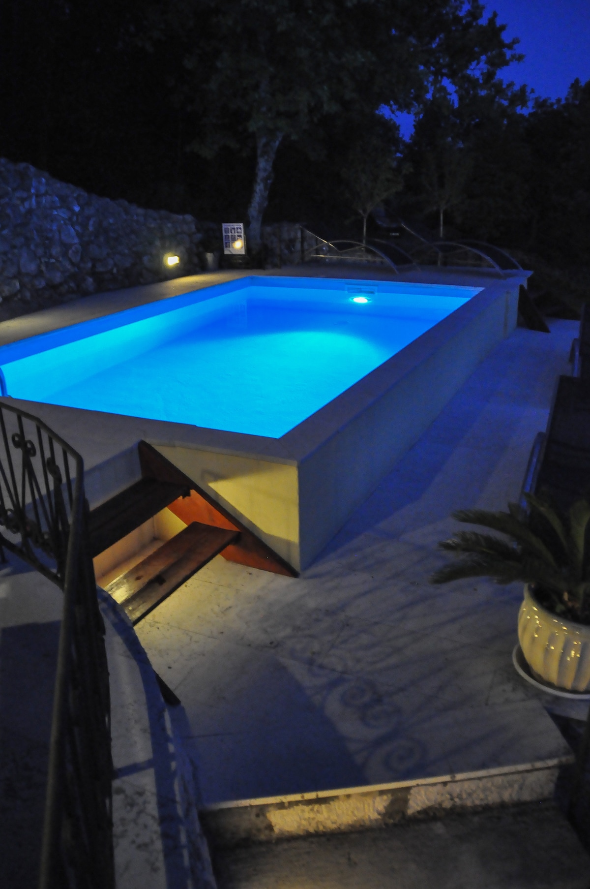 An illuminated pool glows in calming blue tones, surrounded by a stone wall and lush trees. Stairs lead down into the water, with ambient lighting enhancing the tranquil evening setting. Comfortable lounge chairs are positioned nearby for relaxation.