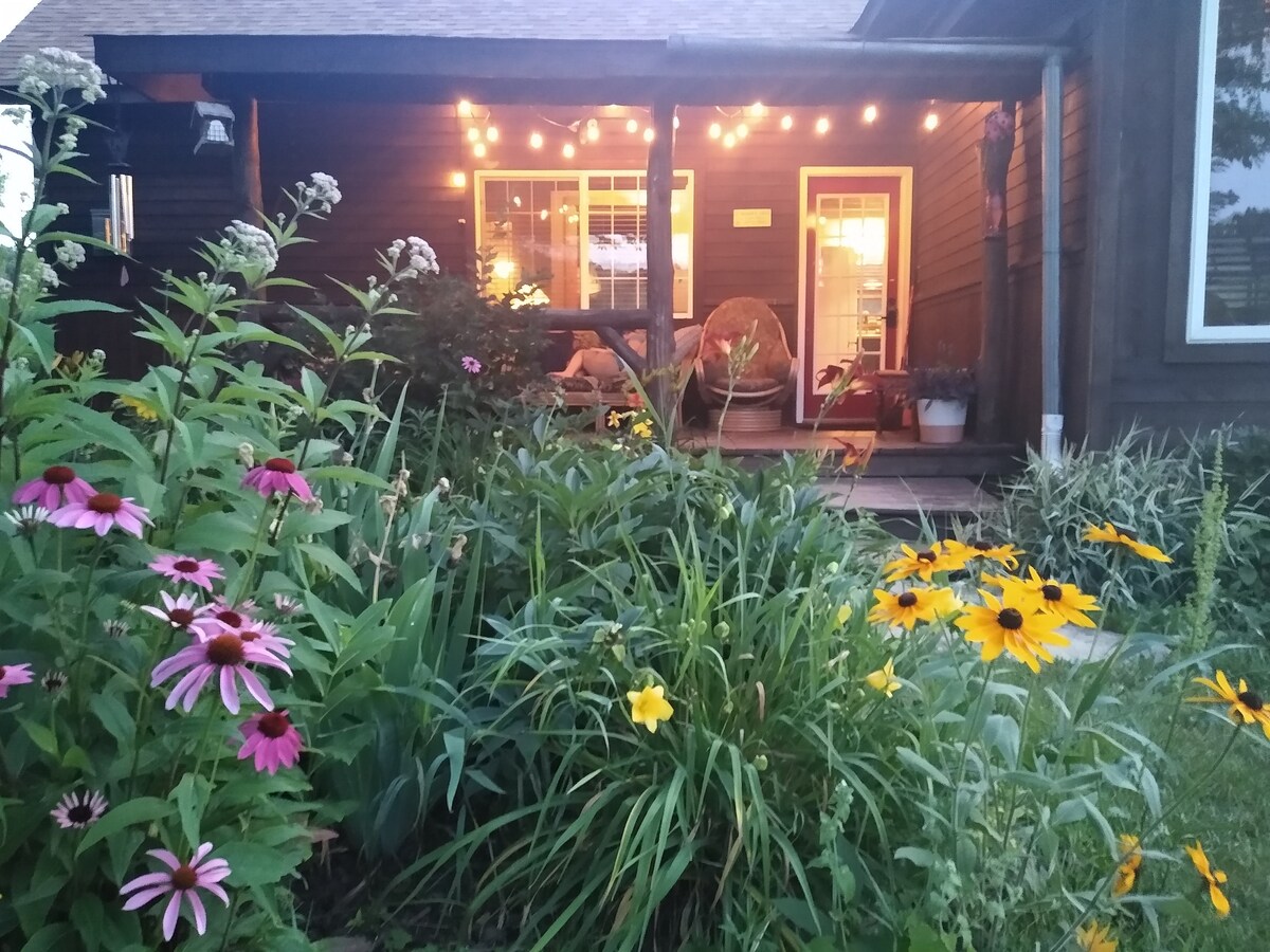 A warm outdoor area is accented with colorful flower beds featuring pink, yellow, and orange blooms. Soft lighting hangs above the porch, guiding the view towards the inviting cabin entrance visible through the partially open door.