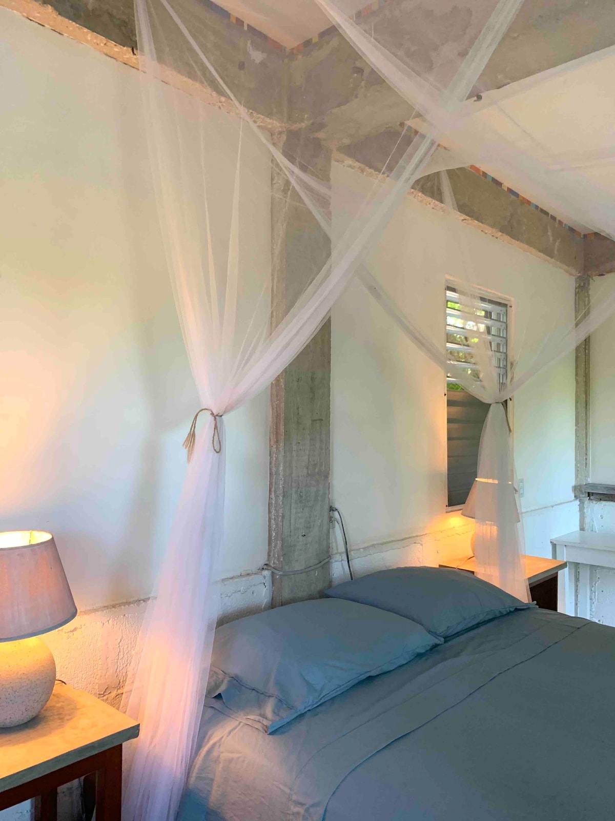 A serene sleeping area is defined by soft gray bedding, complemented by two bedside tables with lamps. Delicate sheer drapes are hung above the bed, filtering warm light from adjacent windows, which provide a sense of openness and tranquility.