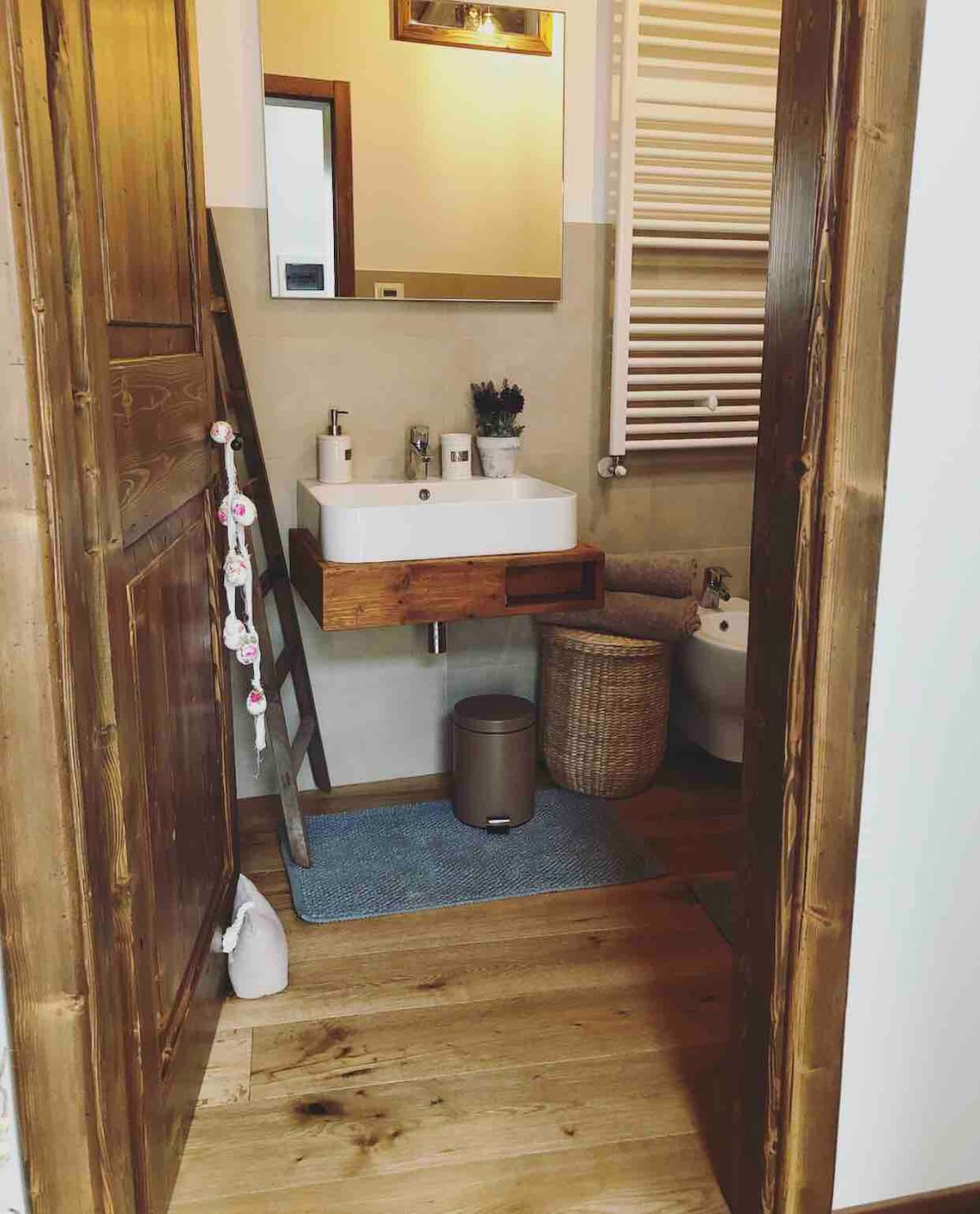 A modern bathroom features a compact sink with wooden accents and a large mirror above. Natural light enters through a wall window. A woven basket is positioned next to the toilet. The warm wooden flooring complements the neutral-toned walls.