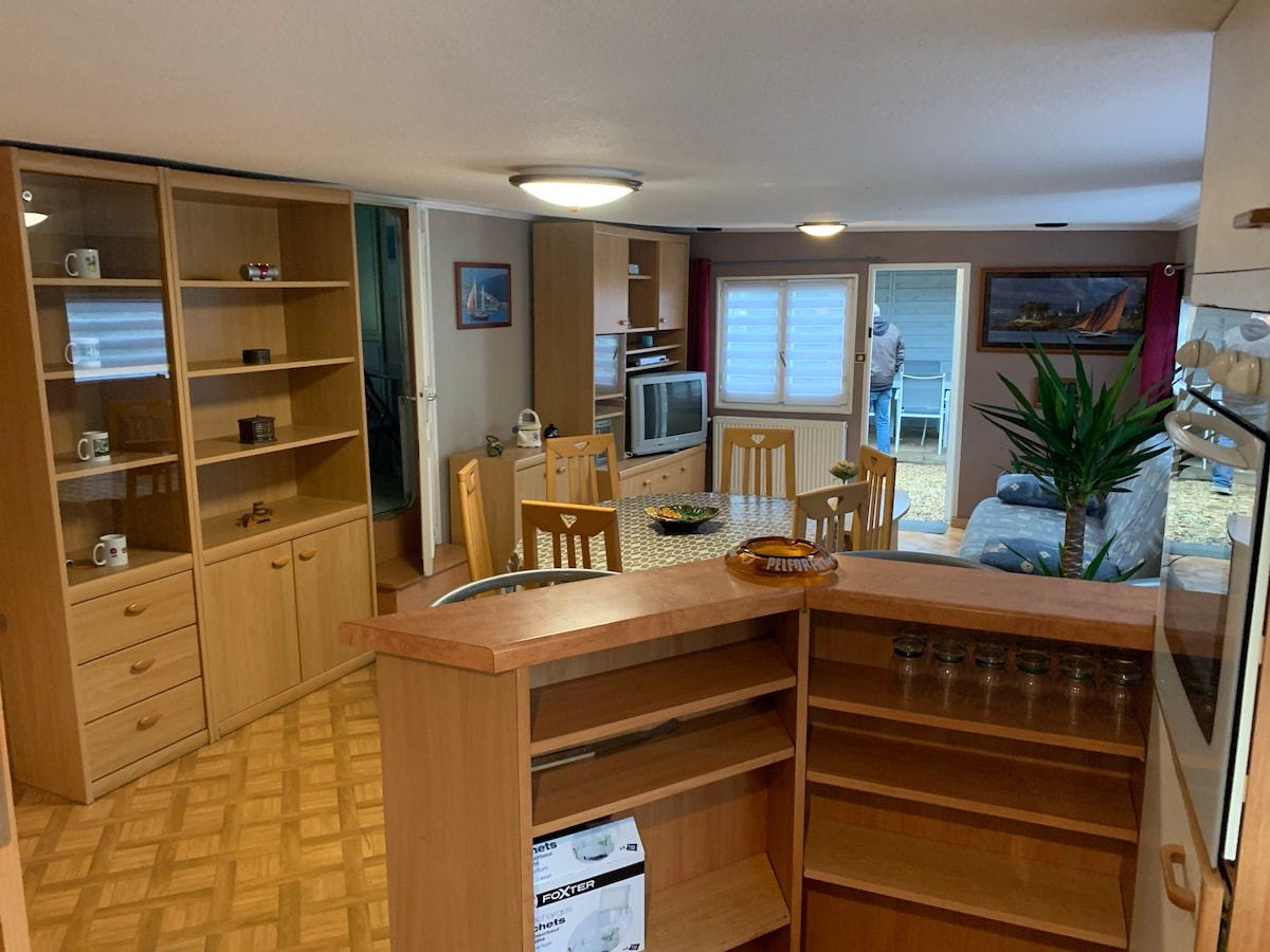 An open living area is showcased, featuring a wooden dining set surrounded by windows that allow natural light to fill the space. Shelves are neatly arranged along the walls, and a cozy seating area with a couch is visible in the background.