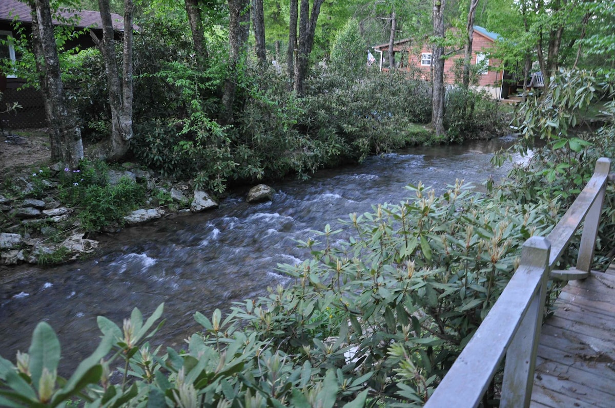 A gentle creek flows through a wooded area, flanked by lush greenery and trees. A wooden bridge crosses the creek, offering a peaceful view of the water and surrounding foliage. A cabin is partially visible in the background, adding to the tranquil setting.