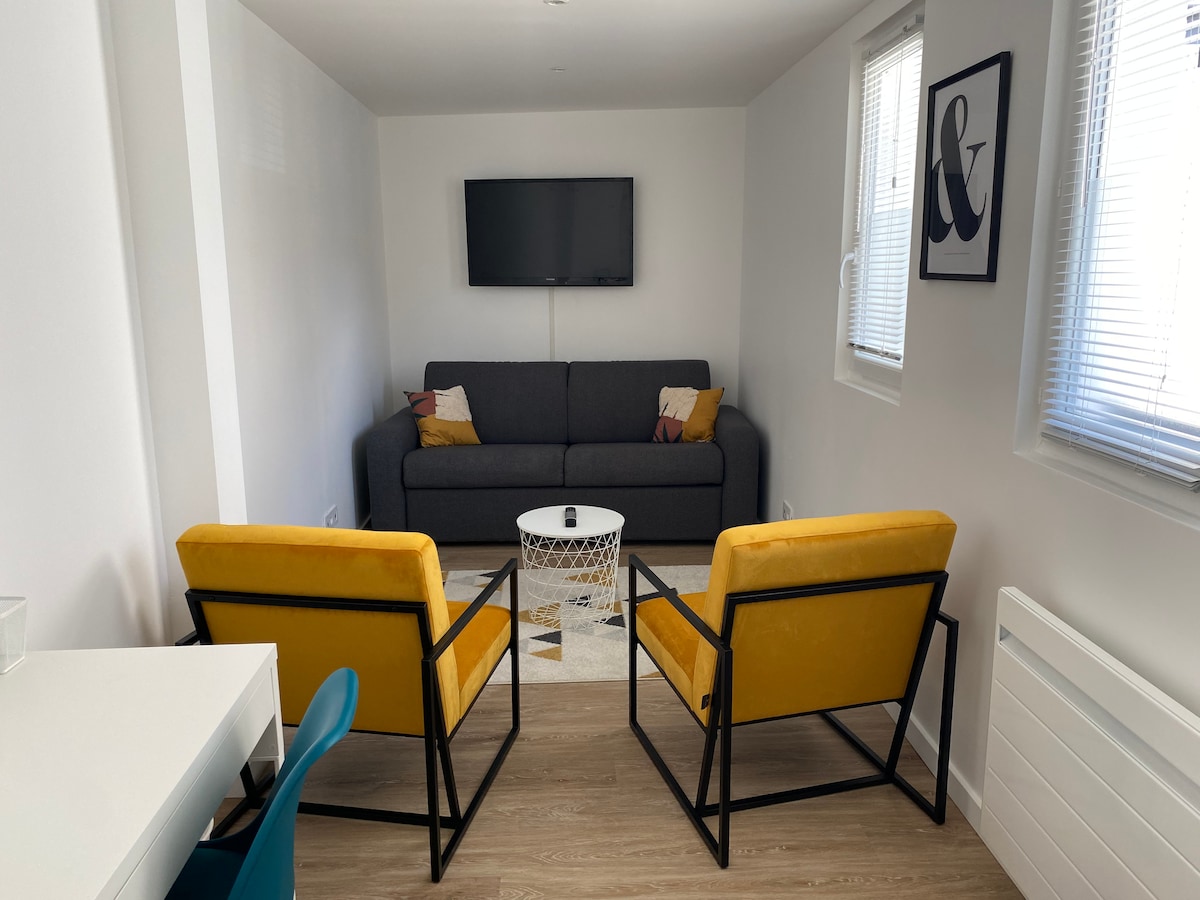 A cozy living area features a gray sofa with decorative pillows and a mounted TV on the wall. Two yellow armchairs are positioned around a round coffee table, placed on a textured rug. Natural light enters through two windows with blinds.