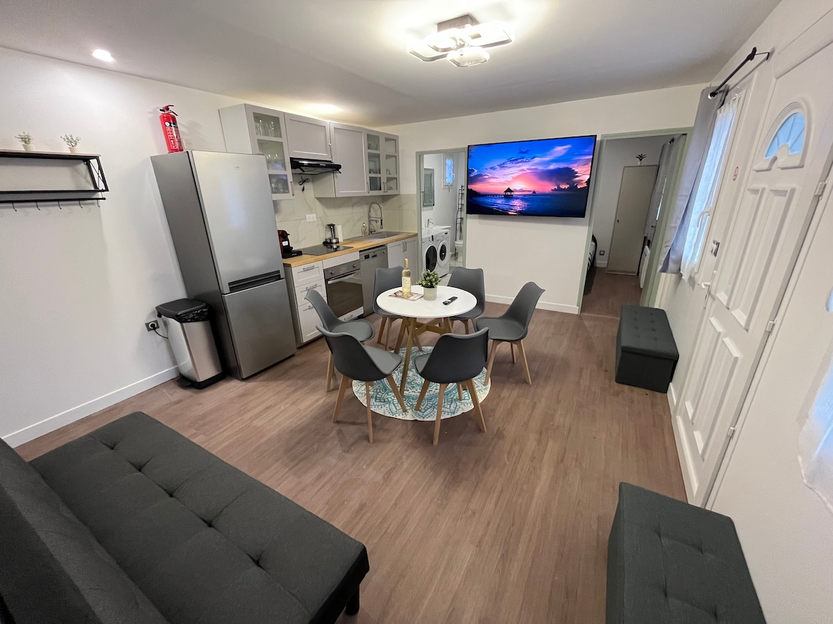 The modern kitchen and living area feature a large television screen displaying a sunset, six dining chairs around a circular table, and a sofa bed. A refrigerator, oven, and washing machine are also present, with natural light coming from the window dressed in light curtains.