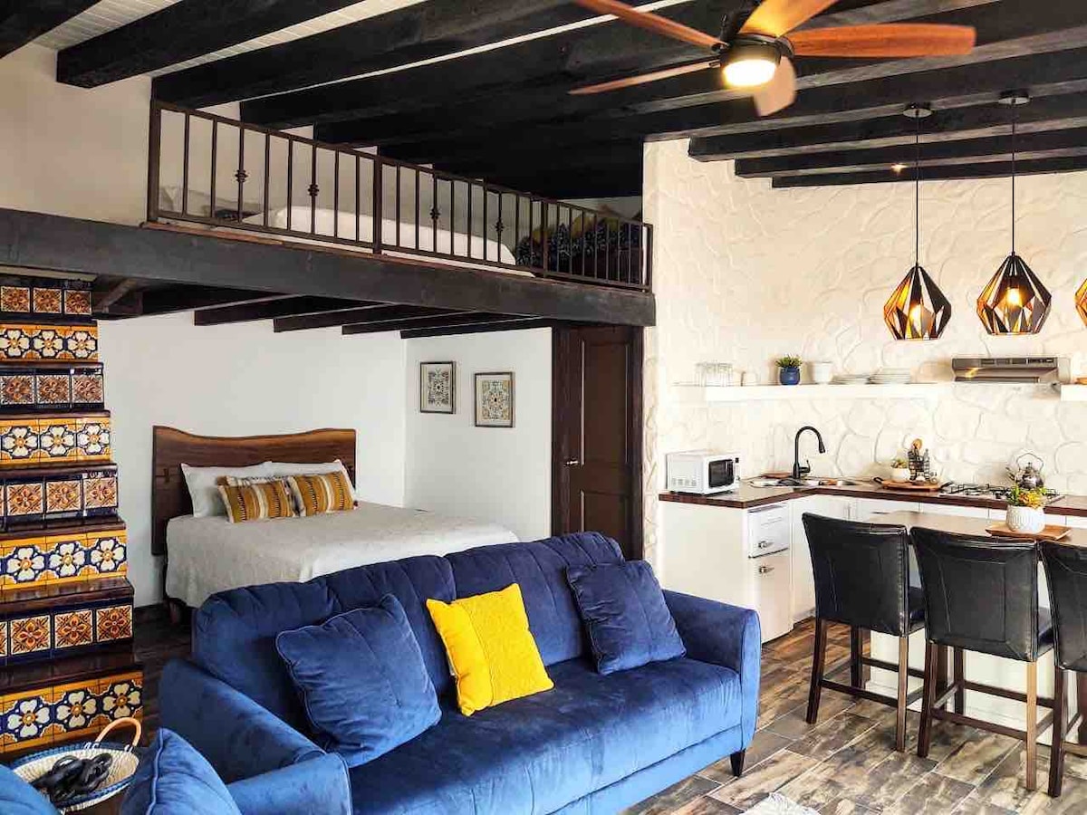 The loft features a spacious living area with a deep blue sofa accented by a yellow pillow. A queen bed sits adjacent to the inviting kitchen, which is illuminated by unique pendant lights. The open design showcases exposed wooden beams and colorful tile detailing.