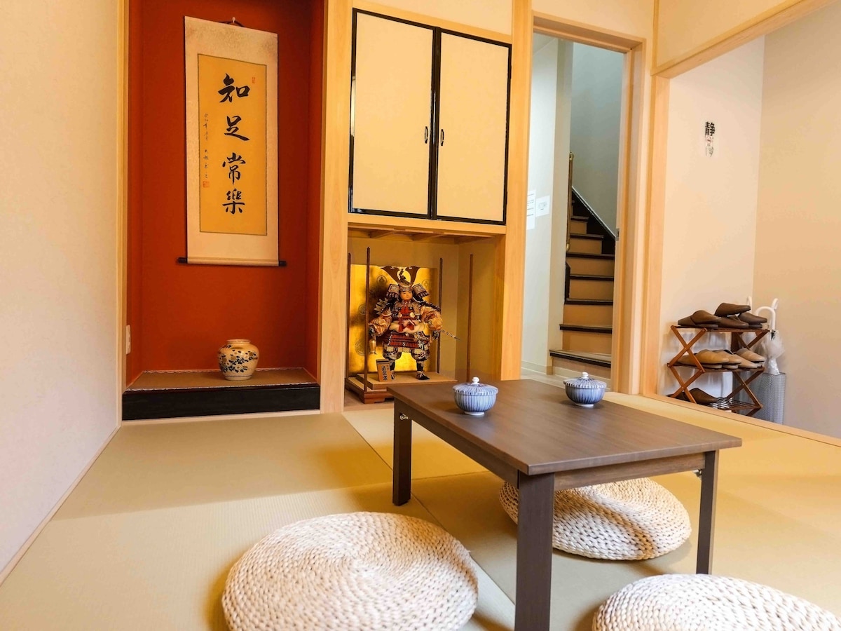 A serene living area is presented with traditional tatami flooring. A low wooden table is set with two small blue bowls. Decorative elements include a calligraphy scroll and a colorful ornamental display, enhancing the cultural ambiance. A staircase is visible in the background.