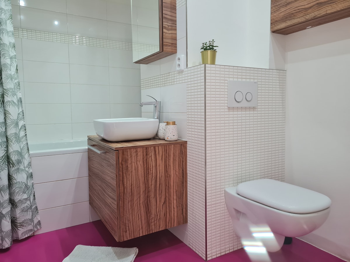 The bathroom features a modern design with a vanity unit made of warm wood, a large basin, and a framed mirror above. A white toilet and a shower curtain with green foliage patterns are visible. Bright pink flooring adds a unique touch to the space.