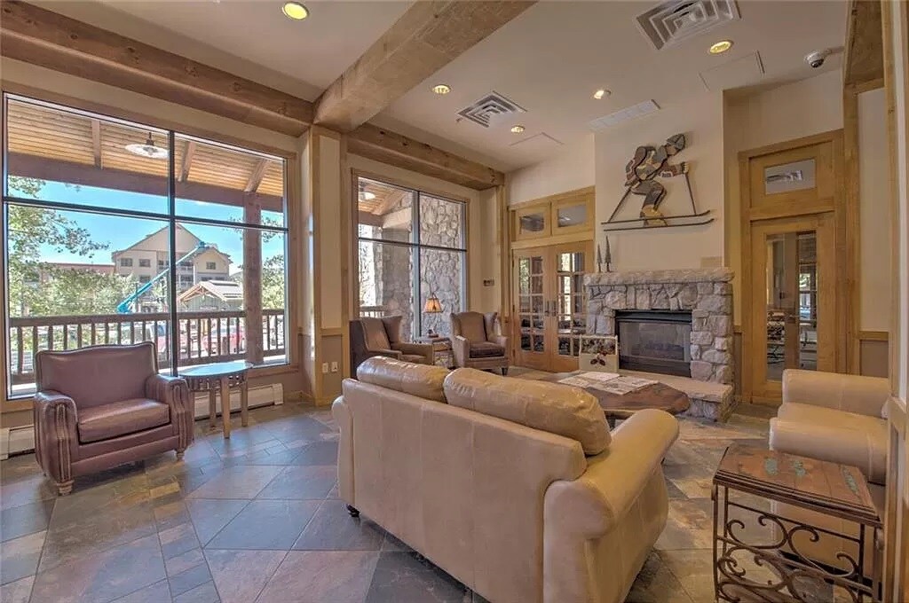 A welcoming lobby area features a stone fireplace surrounded by comfortable seating. Large windows allow natural light to fill the space, showcasing views of the outside. Natural wood beams accent the ceiling, enhancing the cozy ambiance.