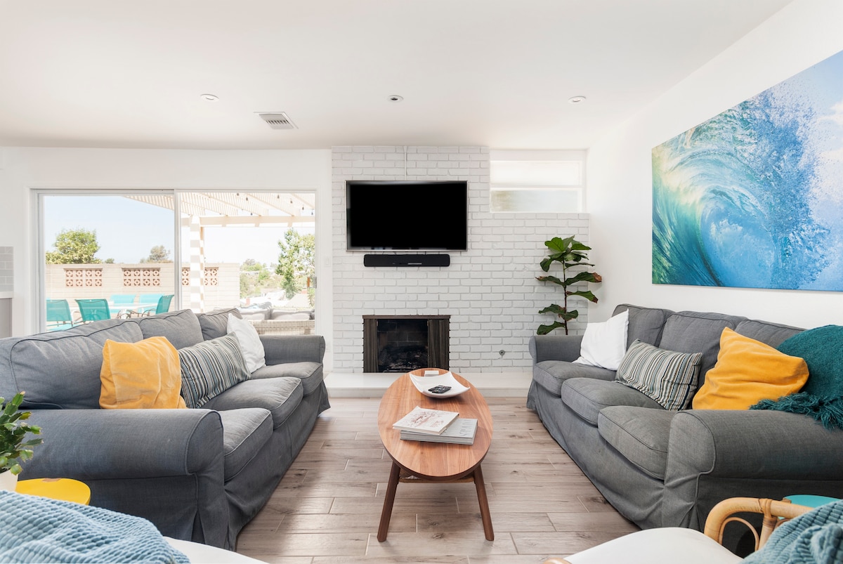 A welcoming living area is furnished with two comfortable gray sofas adorned with colorful throw pillows. A wooden coffee table sits in the center, and a large wall-mounted television is positioned above a white brick fireplace. Natural light flows in through sliding glass doors leading to the outdoor space.