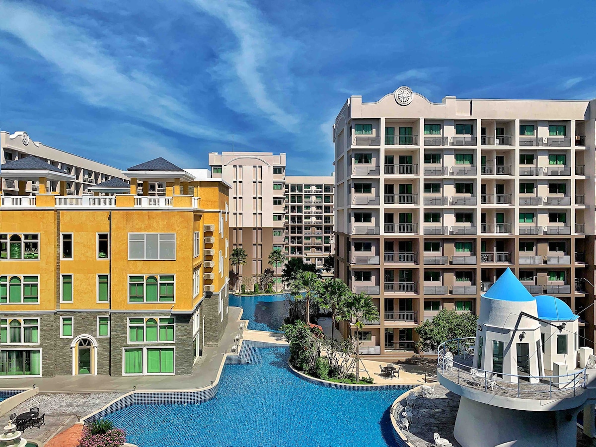 A view of a modern residential complex featuring several buildings surrounding a large swimming pool. Lush landscaping is visible, with palm trees and seating areas enhancing the outdoor space. The architecture includes a mix of colors and styles, creating an inviting atmosphere.