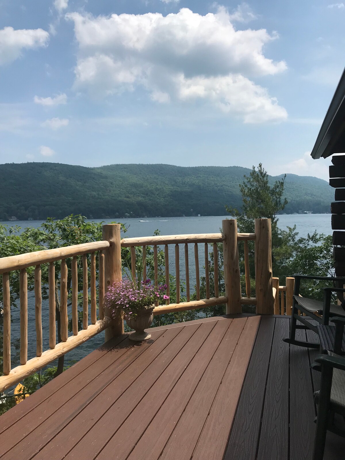 A wooden deck overlooks serene waters and distant mountains. Natural wood railings frame the view, and a potted plant adds a touch of greenery. The sunlit atmosphere enhances the sense of tranquility offered by the lake setting.