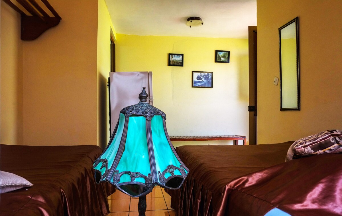 A room features two beds with chocolate-colored linens, providing a cozy sleeping area. A vintage teal lamp adds a pop of color, while the warm yellow walls create an inviting ambiance. A mirror and framed pictures are visible in the background, enhancing the space's charm.