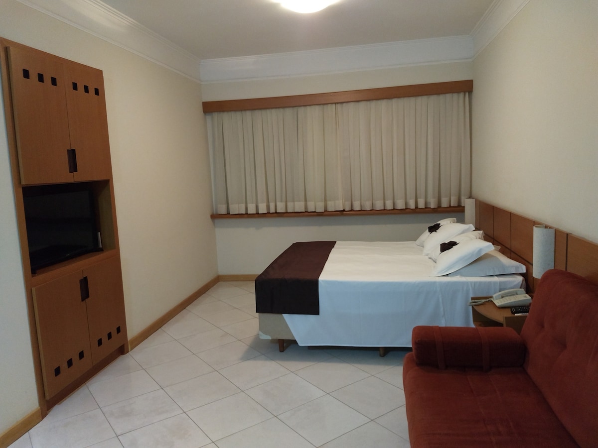 A spacious room features a neatly made bed with a dark headboard and contrasting white linens. A wooden cabinet with an integrated television is visible, along with a brown sofa positioned against the wall. Natural light filters through the sheer curtains covering the window.