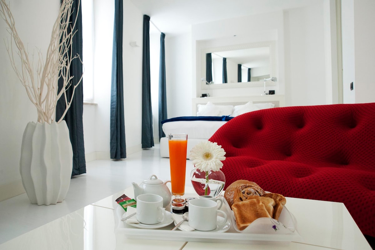 A modern seating area features a bright red sofa with textured upholstery and a low table, showcasing a tray of breakfast items. A glass of orange juice, two cups, pastries, and a floral arrangement are displayed. Natural light enters through large windows, complemented by dark curtains.