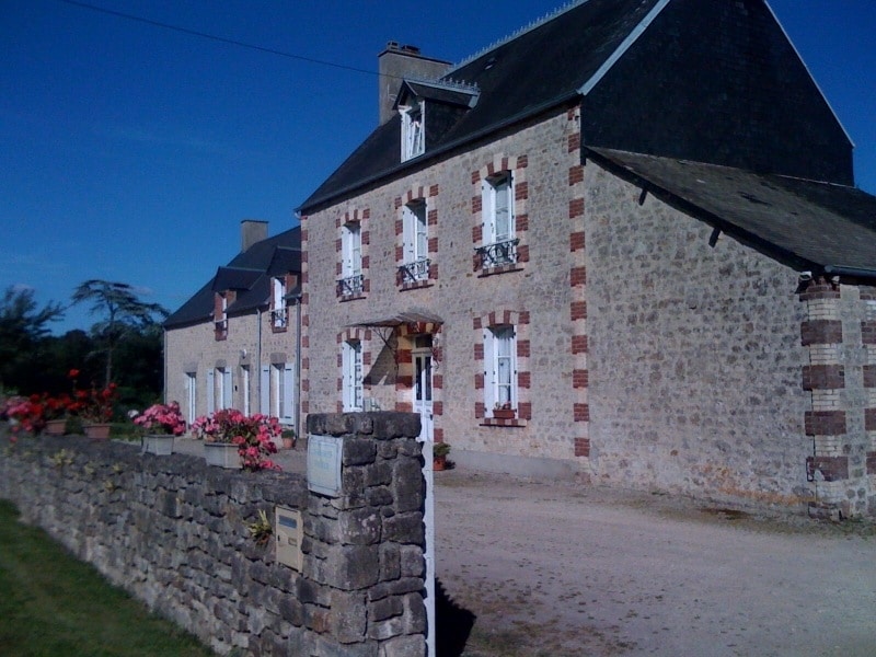A charming rural house features a façade of stone with red brick accents, framed by neatly lined flower beds. The two-story structure is complemented by white window shutters and decorative balconies, while a gravel pathway leads to the entrance. A clear blue sky serves as the backdrop.