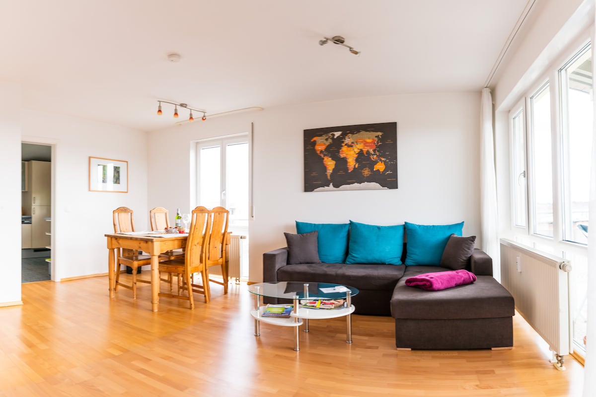 A bright living space features a comfortable dark couch with teal cushions and a small glass table. A wooden dining table with six chairs is positioned near windows, allowing natural light. A world map hangs on the wall, adding interest to the neutral decor.