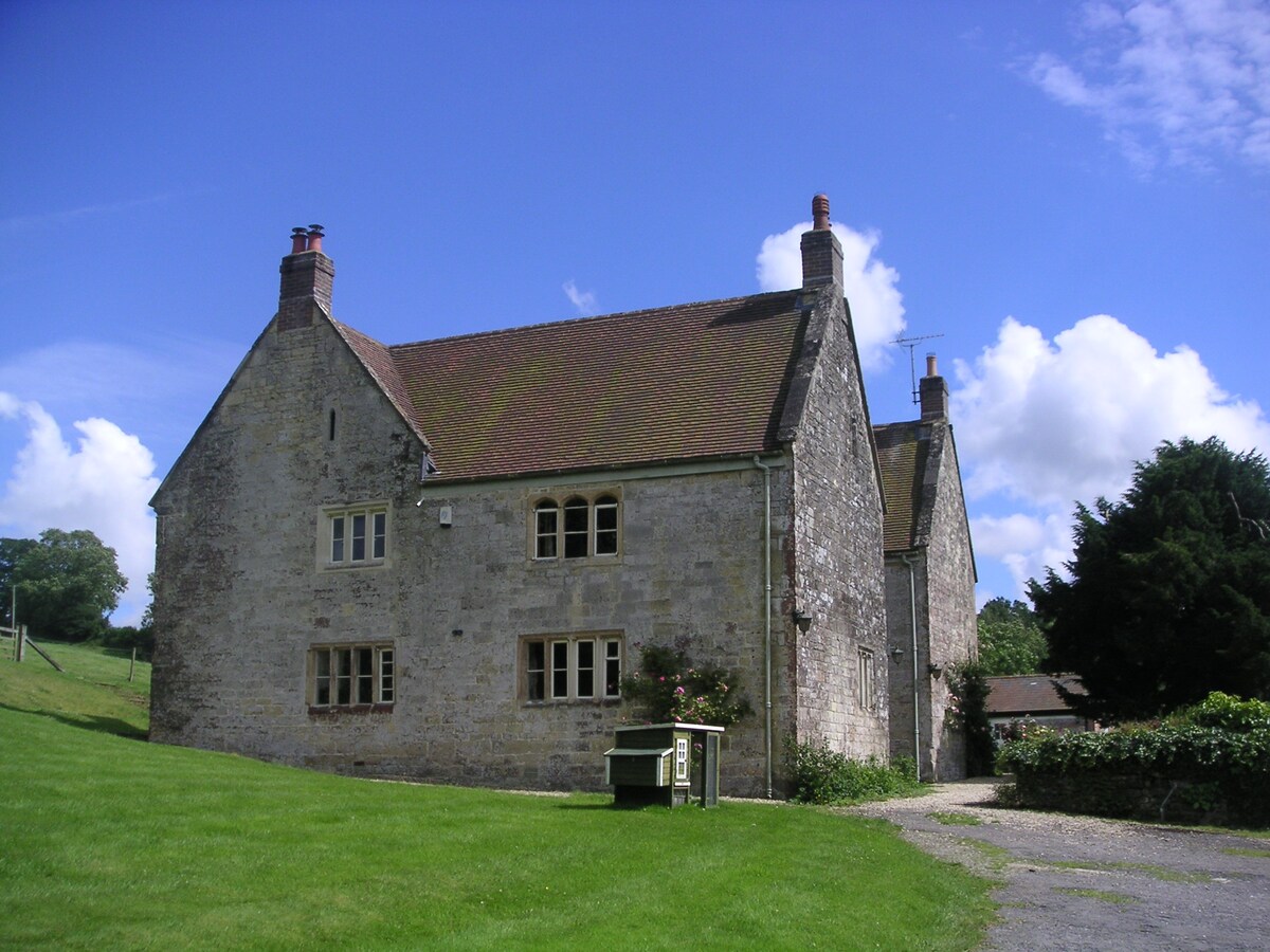 A traditional stone farmhouse sits on a grassy expanse, featuring a sloped roof and multiple chimneys. Large windows allow for ample natural light. The surrounding landscape includes gentle hills and patches of trees, creating a serene countryside setting.